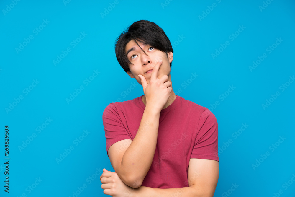 Asian man with red shirt over isolated blue wall standing and thinking an idea Asian man with red shirt over isolated blue wall standing and thinking an idea