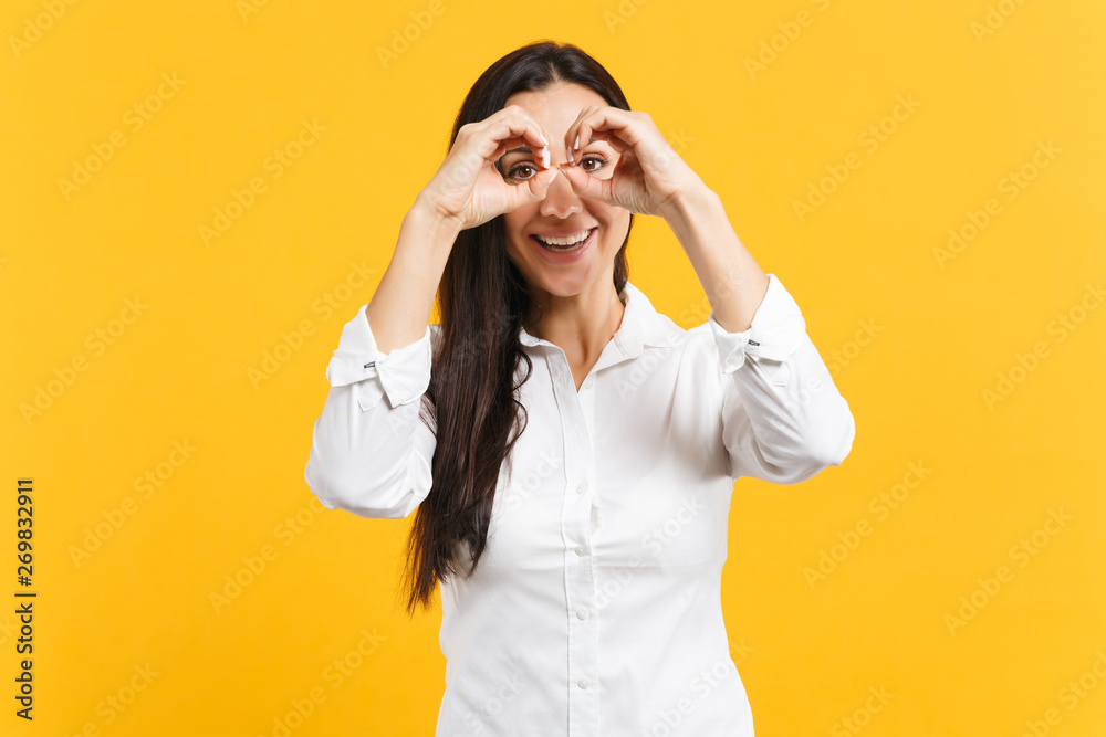 Portrait of funny young woman in white casual shirt holding hands near eyes, imitating glasses or binoculars isolated on yellow orange wall background. People lifestyle concept. Mock up copy space.