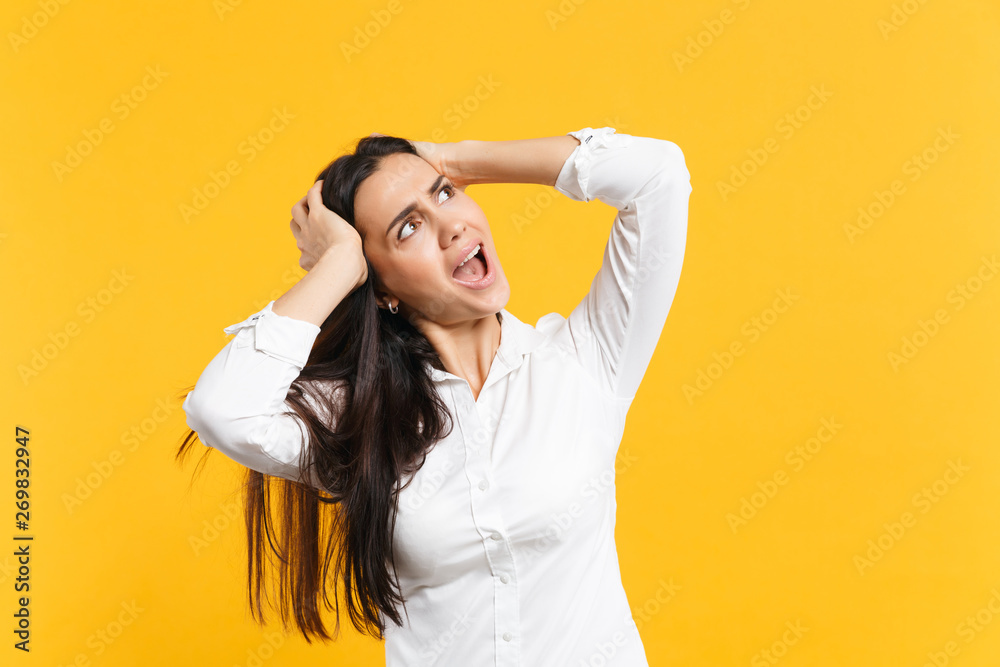 Portrait of irritated angry young woman in white casual shirt looking aside, putting hands on head isolated on yellow orange wall background in studio. People lifestyle concept. Mock up copy space. Portrait of irritated angry young woman in white casual shirt looking aside, putting hands on head isolated on yellow orange wall background in studio. People lifestyle concept. Mock up copy space.