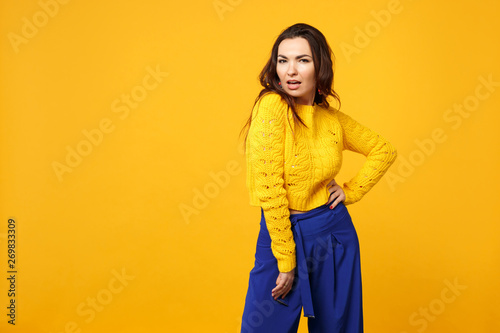 Portrait of passionate young woman in sweater, blue trousers standing with arm akimbo on waist isolated on yellow orange wall background. People sincere emotions lifestyle concept. Mock up copy space.