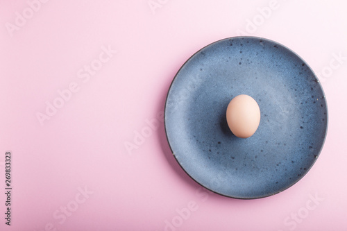 Raw uncooked single egg on a blue ceramic plate on a pink pastel background. top view.