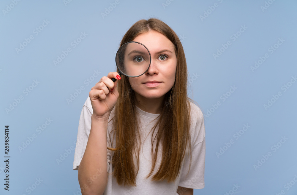 Young woman over blue wall taking a magnifying glass and looking through it Young woman over blue wall taking a magnifying glass and looking through it