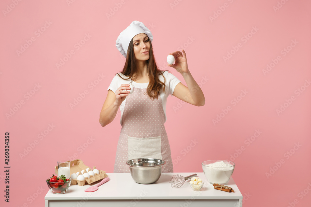 Female chef cook confectioner or baker in apron white t-shirt, toque chefs hat cooking cake or cupcake at table hold eggs isolated on pink pastel background in studio. Mock up copy space food concept. Female chef cook confectioner or baker in apron white t-shirt, toque chefs hat cooking cake or cupcake at table hold eggs isolated on pink pastel background in studio. Mock up copy space food concept.