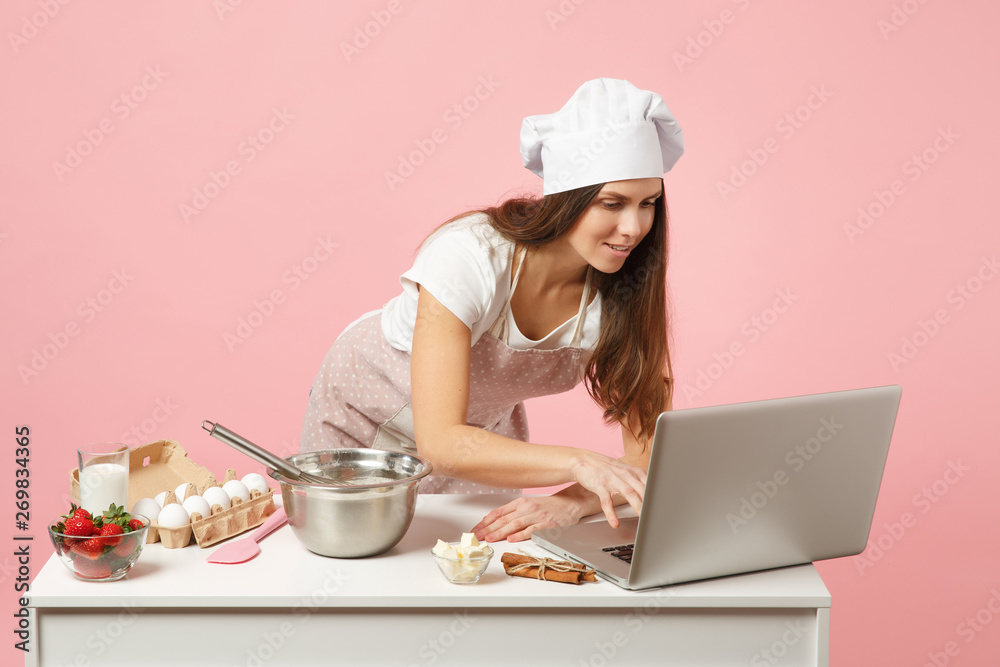 Chef cook confectioner or baker in apron white t-shirt, toque chefs hat cooking Christmas cake or cupcake at table looking for recipe on pc laptop isolated on pink pastel background. Mock up concept. Chef cook confectioner or baker in apron white t-shirt, toque chefs hat cooking Christmas cake or cupcake at table looking for recipe on pc laptop isolated on pink pastel background. Mock up concept.