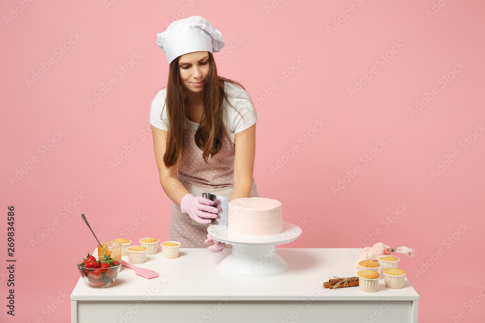Chef cook confectioner or baker in white t-shirt toque chefs hat cooking at table isolated on pink pastel background in studio. Cream application, cake making process. Mock up copy space food concept. Chef cook confectioner or baker in white t-shirt toque chefs hat cooking at table isolated on pink pastel background in studio. Cream application, cake making process. Mock up copy space food concept.