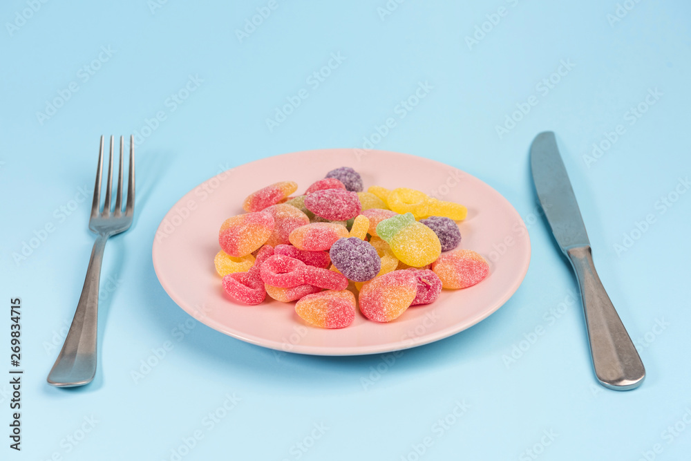 Fork, knife and pink plate with gummy candies isolated on blue background Fork, knife and pink plate with gummy candies isolated on blue background