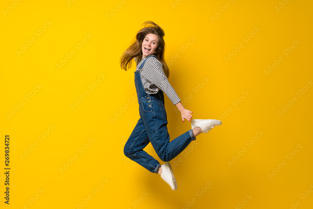 Young girl with overalls jumping over isolated yellow background