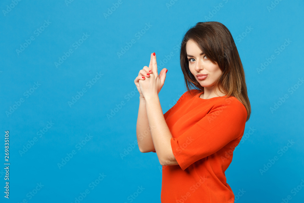 Beautiful woman wearing orange dress looking camera holding hands like gun, side profile view isolated over blue wall background, studio portrait. People lifestyle fashion concept. Mock up copy space.