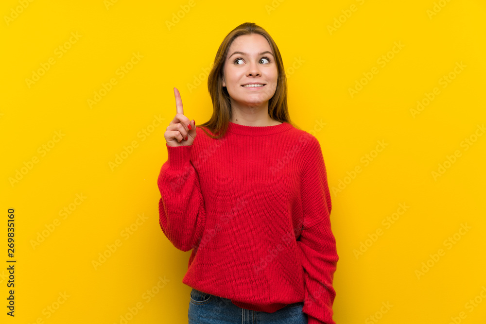 Young woman over yellow wall intending to realizes the solution while lifting a finger up Young woman over yellow wall intending to realizes the solution while lifting a finger up