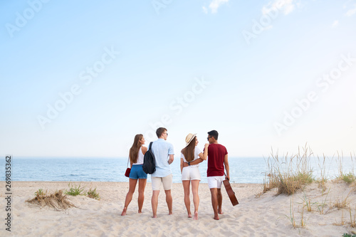 Group of friends going to sandy beach at sunset. Young men and women walking to the shore to relax near sea and play guitar. Teenagers on holidays. Boyfriend and girlfriend embrace. Isolated view.