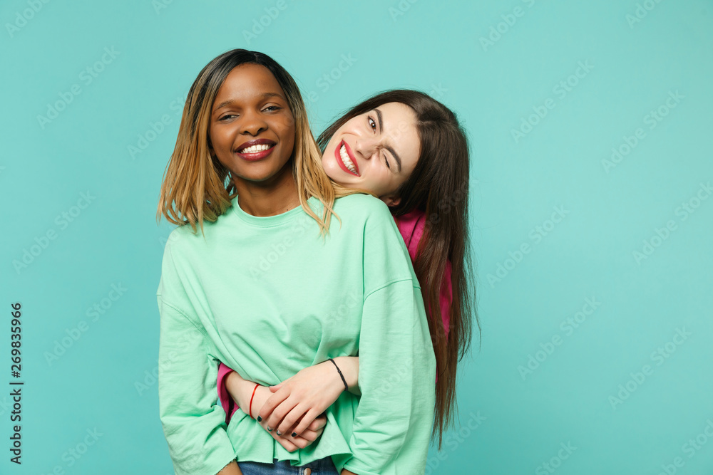 Two young women friends european and african american in pink green clothes standing posing isolated on blue turquoise wall background, studio portrait. People lifestyle concept. Mock up copy space. Two young women friends european and african american in pink green clothes standing posing isolated on blue turquoise wall background, studio portrait. People lifestyle concept. Mock up copy space.