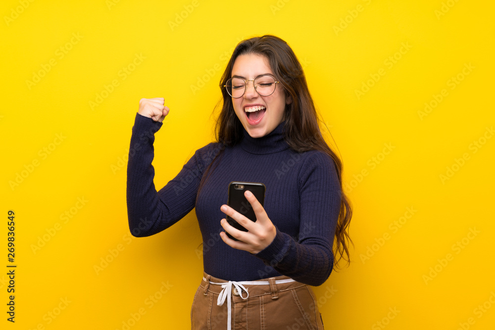 Teenager girl over isolated yellow wall with phone in victory position Teenager girl over isolated yellow wall with phone in victory position