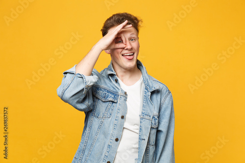 Cheerful young man in denim clothes showing tongue, holding hand near eyes, imitating glasses or binoculars isolated on yellow orange background. People sincere emotions concept. Mock up copy space.