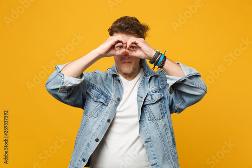 Portrait of young man in denim casual clothes hold hands near eyes imitating glasses or binoculars isolated on yellow orange background. People sincere emotions lifestyle concept. Mock up copy space.