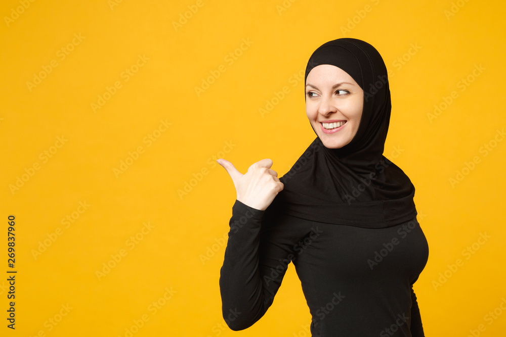 Smiling young arabian muslim woman in hijab black clothes showing pointing copy space with hands finger isolated on yellow wall background, studio portrait. People religious lifestyle concept. Mock up Smiling young arabian muslim woman in hijab black clothes showing pointing copy space with hands finger isolated on yellow wall background, studio portrait. People religious lifestyle concept. Mock up