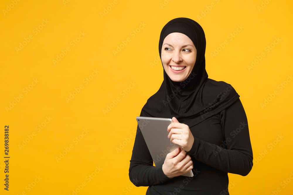 Young arabian muslim employee woman in hijab black clothes hold and work tablet pc computer isolated on yellow wall background, studio portrait. People religious lifestyle concept. Mock up copy space. Young arabian muslim employee woman in hijab black clothes hold and work tablet pc computer isolated on yellow wall background, studio portrait. People religious lifestyle concept. Mock up copy space.