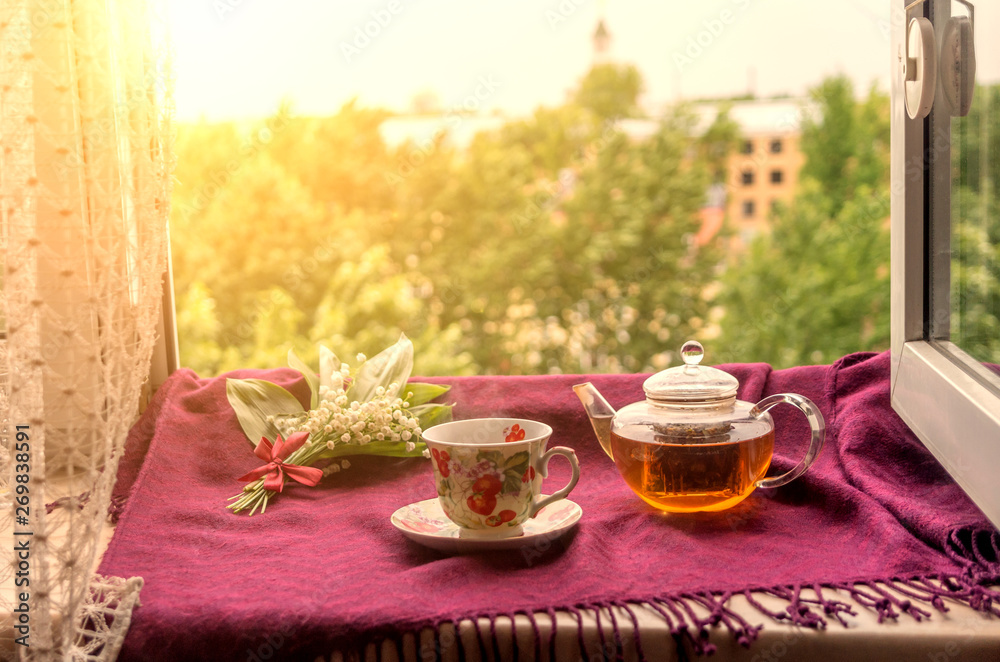 Tea pot, cup, saucer and a bouquet of spring flowers on a window sill. Enjoy lilies of the valley scent, hot drink and a beautiful view at sunset time  in summer. Golden evening light through window