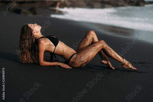Fotografie, Obraz Attractive sexy girl in swimwear rests on black volcanic sand beach