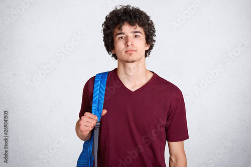 Portrait of thoughtful good looking guy with curly black hair standing straight, having blue backpack on his back, wearing dark red t shirt, being confident and serious, looking directly at camera. photo