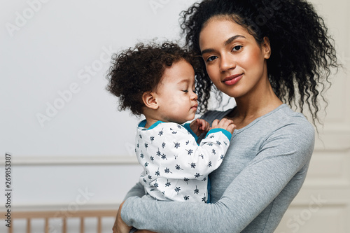 Young mom holding her sleepy baby boy. Beautiful mother with son in living room. photo