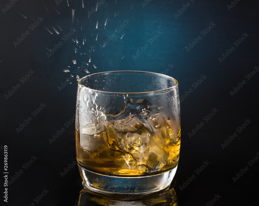 A glass of whiskey with ice on a dark color photo with splashes from falling ice A glass of whiskey with ice on a dark color photo with splashes from falling ice