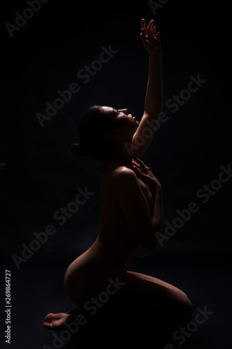 Nude woman kneeling in darkness and reaching up to light photo