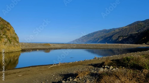 Slow pan of coastal lagoon on calm morning stillness, sand berm and large imposing Big Sur coastline mountains in background