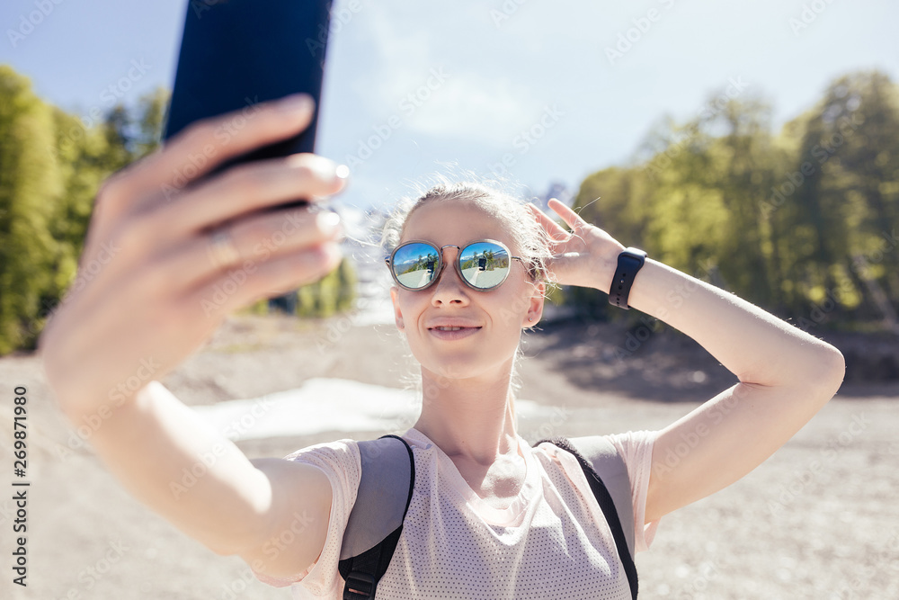 Naklejka premium Beautiful young girl traveler environmentalist makes selfie in the mountains during the sunny summer weather. Concept candid lifestyle and trekking in the mountains