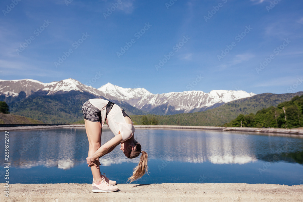 Fit young girl fitness model doing exercises in nature on the background of a large lake mountains and blue sky on a sunny summer day. Concept of sport and workout in nature. Copyspace