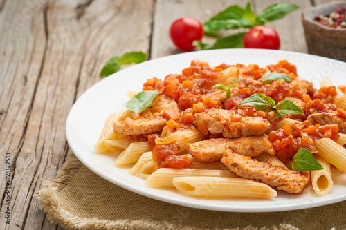 Penne pasta, chicken or turkey fillet, tomato sauce with basil leaves on old rustic wooden background. Copy space, side view