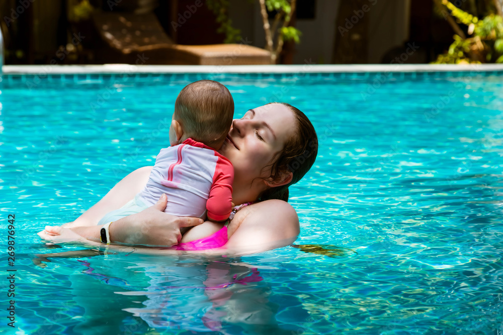 Happy mother with little baby daughter swims in the pool at summer holiday. Sunny day. Tropics. Infant watching around