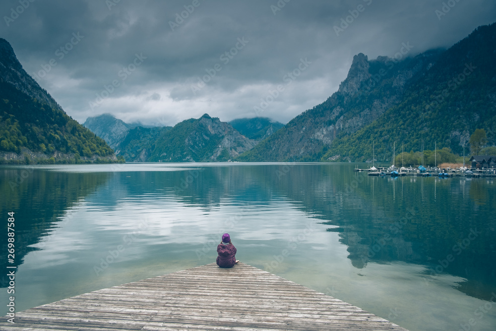 Kobieta samotnie siedzi na pomoście nad górskim jeziorem w pochmurny dzień	 - obrazy, fototapety, plakaty 