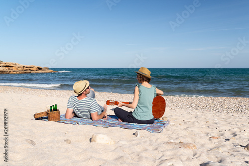 Young couple having a romantic picnic while the woman is playing the guitar on a mediterranean beach at sunset. Music and love
