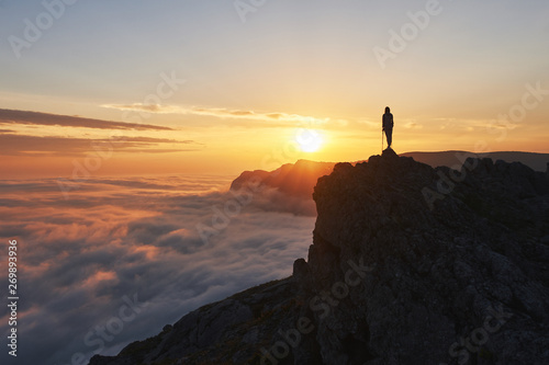 silhouette of hiker woman with trekking poles stands at rock and looks at aerial view in the mountains above the clouds. Mountains sunset landscape with tourist silhouette in orange sun light.