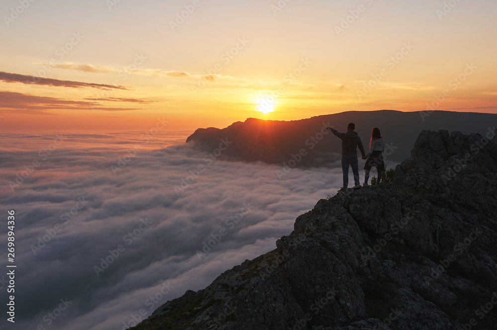 Young couple of hikers stands in the mountains above the clouds and looks at beautiful sunset view. Man and woman hold hands silhouette. Family tourism concept. Love in the mountains