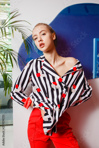 Fashion young girl blogger dressed in stylish striped shirt and red trousers poses on the background of white and blue wall
