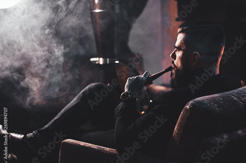 Groomed bearded man is relaxing on lounge near fireplace while smoking hookah. He has tattoo on his hand. photo