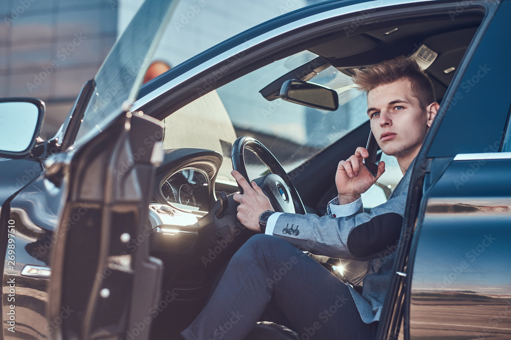 Young attractive businessman is sitting in his car at the parking while talking by mobile phone.