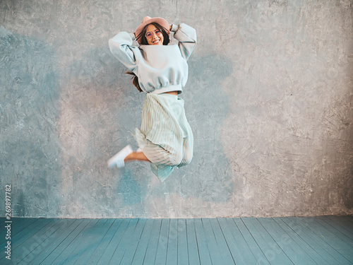 Beautiful cute smiling model jumping. Girl in summer hipster hoodie and skirt. Model having fun and going crazy near gray wall in studio