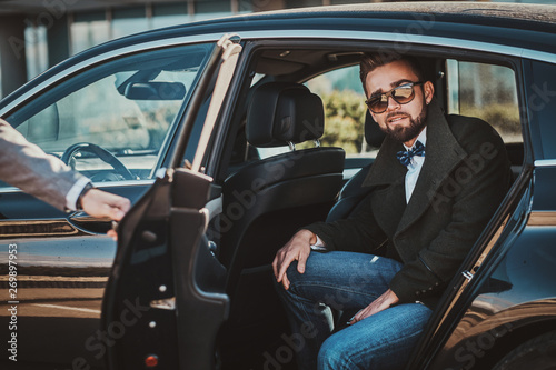 Busy nice businessman in sunglasses is sitting in the car while his elegant assistant is opening door for him.