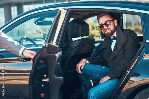 Busy nice businessman in sunglasses is sitting in the car while his elegant assistant is opening door for him. © Fxquadro
