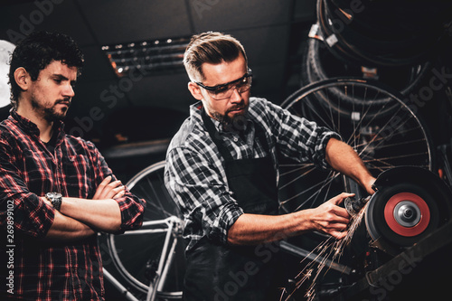 Skilled repairmaster is working with machine tool at busy bucycle workshop and his collegue is standing nearby. photo