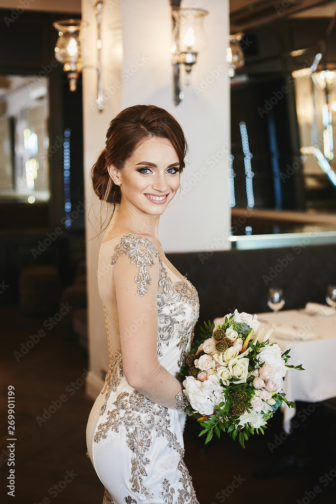 Naklejka premium Beautiful brunette model girl with wedding hairstyle and blue eyes in long fashionable evening dress holding the bouquet of fresh flowers and posing in a cafe interior
