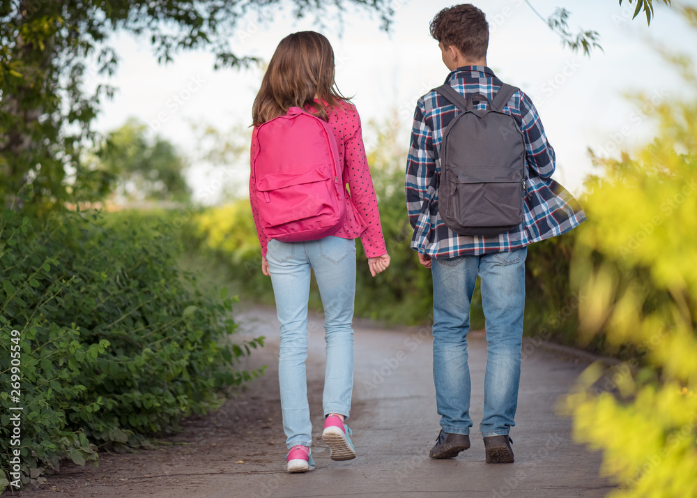 Back view - two schoolchildren go to school with backpacks. Cute children - teen girl and boy with bags Back to school. Concepts of friends, childhood and education.
