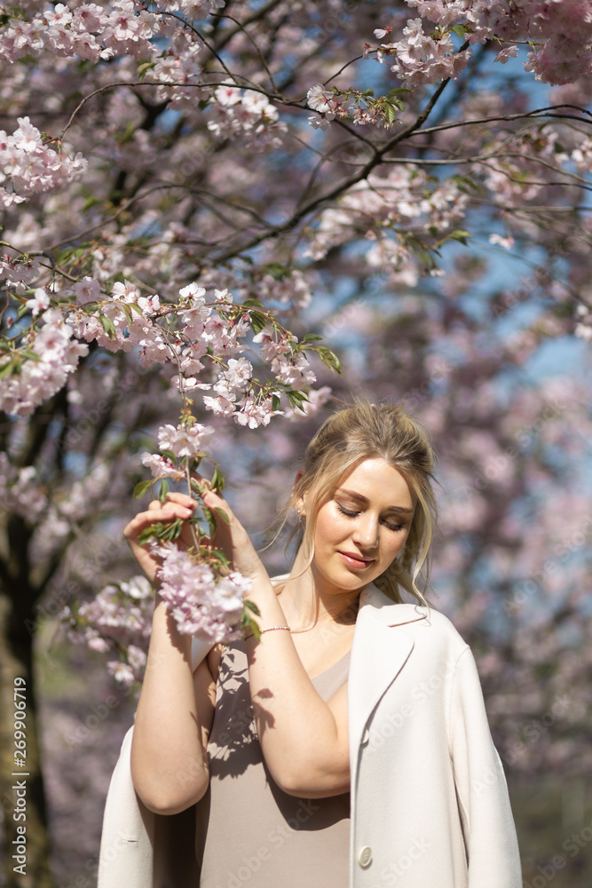 Beautiful blonde young woman in Sakura Cherry Blossom park in Spring enjoying nature and free time during her traveling tourist free time - Wearing white pants and t-shirt with a beige jacket