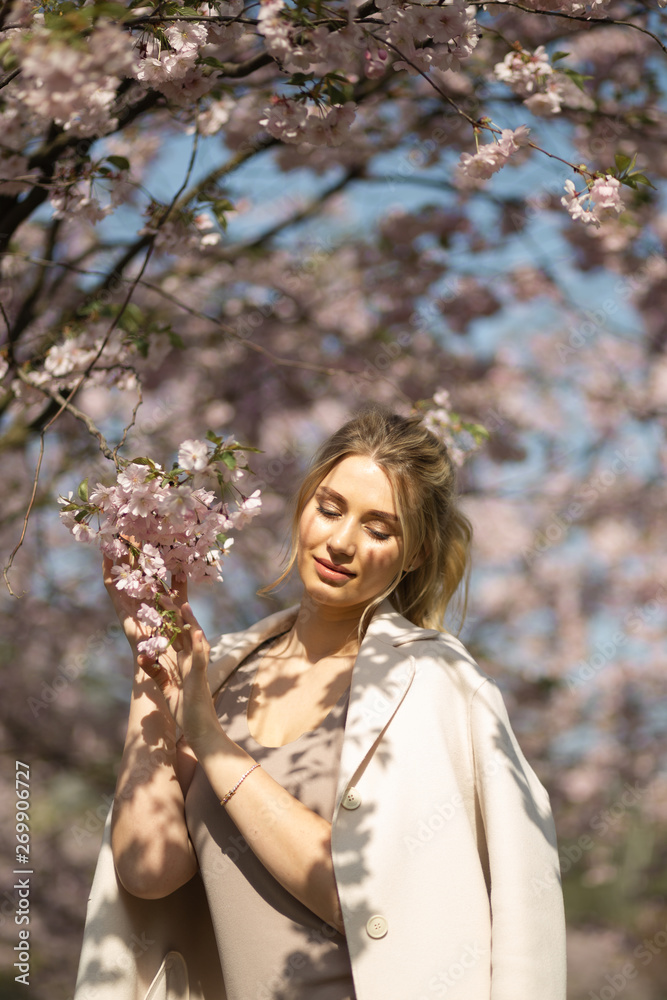 Beautiful blonde young woman in Sakura Cherry Blossom park in Spring enjoying nature and free time during her traveling tourist free time - Wearing white pants and t-shirt with a beige jacket Beautiful blonde young woman in Sakura Cherry Blossom park in Spring enjoying nature and free time during her traveling tourist free time - Wearing white pants and t-shirt with a beige jacket