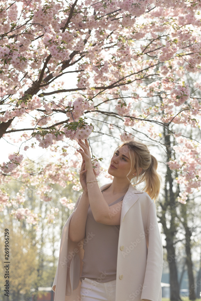 Beautiful blonde young woman in Sakura Cherry Blossom park in Spring enjoying nature and free time during her traveling tourist free time - Wearing white pants and t-shirt with a beige jacket Beautiful blonde young woman in Sakura Cherry Blossom park in Spring enjoying nature and free time during her traveling tourist free time - Wearing white pants and t-shirt with a beige jacket