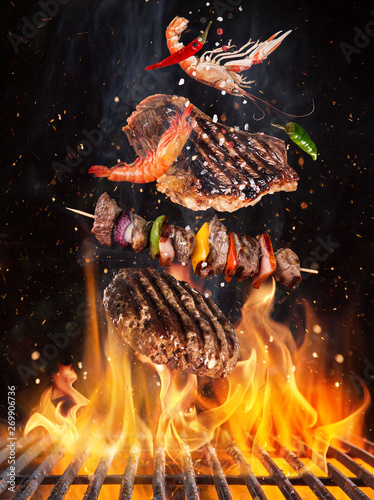 Tasty beef steaks and skewers flying above cast iron grate with fire flames. Freeze motion barbecue concept.