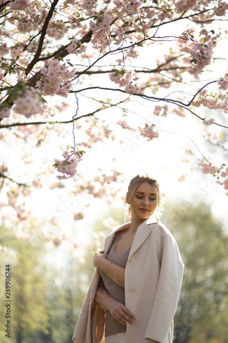 Beautiful blonde young woman in Sakura Cherry Blossom park in Spring enjoying nature and free time during her traveling tourist free time - Wearing white pants and t-shirt with a beige jacket