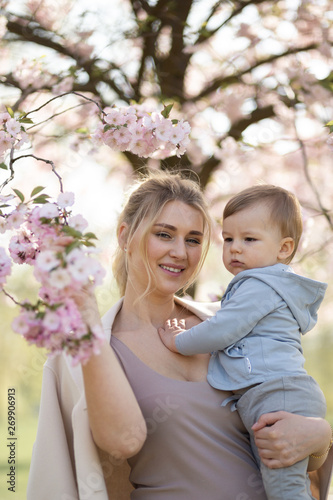 Young mother mom holding her little baby son boy child under blossoming SAKURA Cherry trees with falling pink petals and beautiful flowers